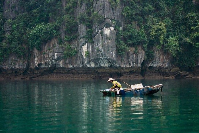 Hình ảnh một ngư dân trên chiếc thuyền đánh cá nhỏ giữa vịnh nước lặng, với những vách đá vôi trầm mặc hiện lên trong sương.